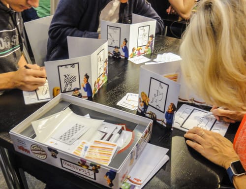 Personas jugando un juego de mesa con hojas de dibujo y cartas en una reseña educativa de Picassimo