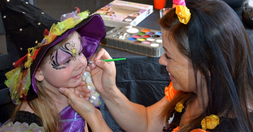 Madre aplicando productos de maquillaje creativo para niños en la cara de su hija disfrazada de bruja
