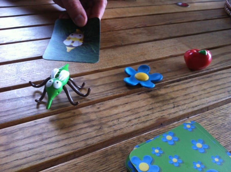 Mano sosteniendo carta del juego Mosquito con piezas de insecto, flor y manzana en mesa de madera. Aprende cómo jugar a Mosquito de Djeco